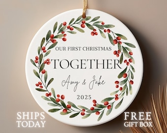 Könnte beinhalten: Weißer Keramik-Weihnachtsschmuck mit goldener Aufhängeschnur. Das Ornament zeigt einen Kranz aus grünen Blättern und roten Beeren um den Text "OUR FIRST CHRISTMAS TOGETHER Amy & Jake 2025."