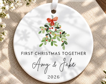 May include: A round white ceramic ornament featuring a watercolour illustration of green mistletoe with a red bow. The text reads FIRST CHRISTMAS TOGETHER, Amy & Jake, and 2026. A white ribbon is attached to the top for hanging purposes.