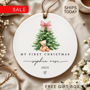 May include: A white ceramic Christmas ornament with a watercolor design of a Christmas tree, teddy bear, and a pink bow. The text reads "My First Christmas" and "Sophia Rose 2025". A brown string is attached for hanging.