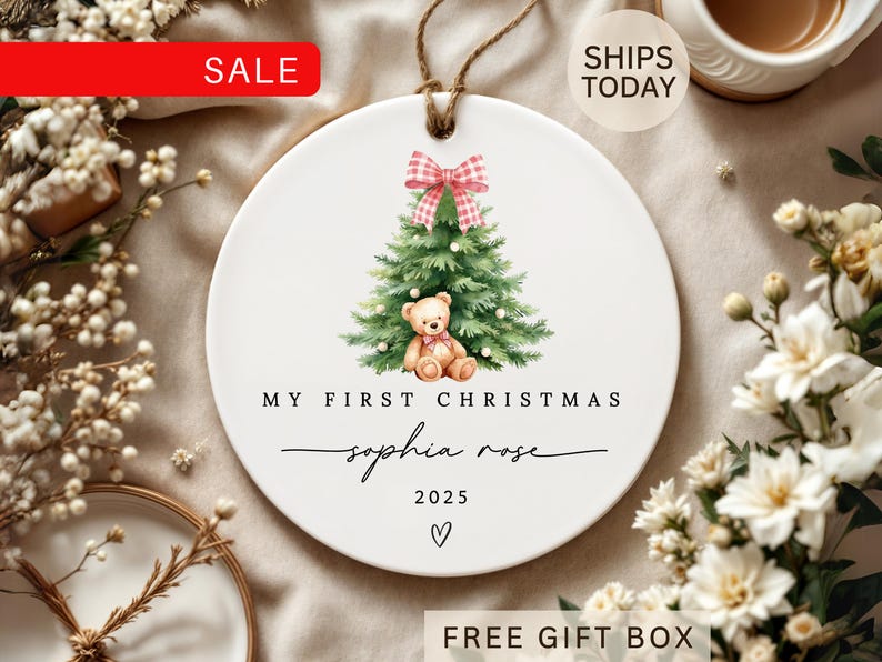 May include: A white ceramic Christmas ornament with a watercolor design of a Christmas tree, teddy bear, and a pink bow. The text reads "My First Christmas" and "Sophia Rose 2025". A brown string is attached for hanging.