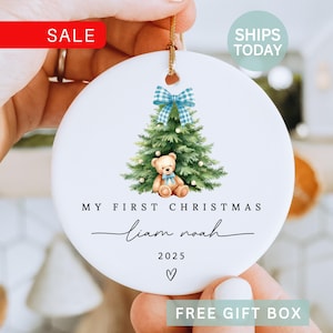 May include: A white ceramic Christmas ornament with a watercolor illustration of a Christmas tree, teddy bear, and a blue plaid bow. The text reads "My First Christmas" and a name, with the year 2025. A gold string is attached for hanging.