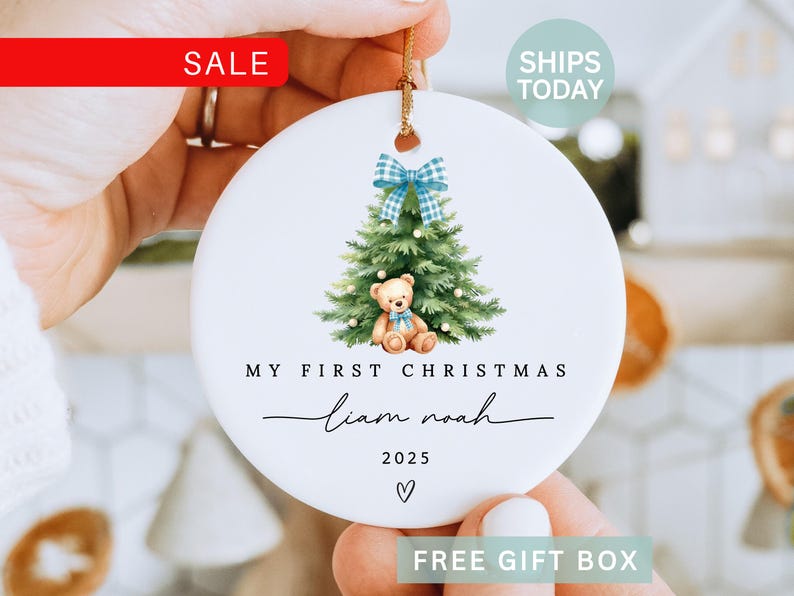 May include: A white ceramic Christmas ornament with a watercolor illustration of a Christmas tree, teddy bear, and a blue plaid bow. The text reads "My First Christmas" and a name, with the year 2025. A gold string is attached for hanging.
