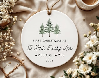 May include: A white ceramic Christmas ornament with a string for hanging. It features three green pine trees and the text "FIRST CHRISTMAS AT 15 Pink Daisy Ave AMELIA & JAMES 2025". The ornament is surrounded by flowers.