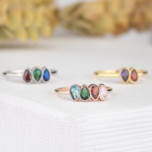 May include: Four rings with pear-shaped gemstones. The rings are in different colors: blue, green, red, and clear. The rings are on a white surface.