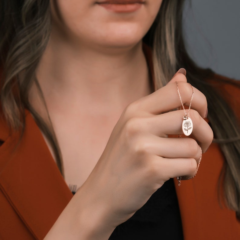 May include: Rose gold necklace featuring an oval pendant with a rose engraving. The pendant hangs from a delicate chain. The person in the image is wearing a rust-colored blazer and a black top.