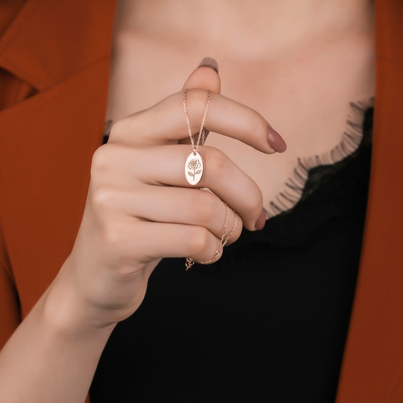 May include: Rose gold necklace featuring a pendant with a rose design. The oval pendant hangs from a delicate chain. The necklace is displayed against a black top and an orange blazer.