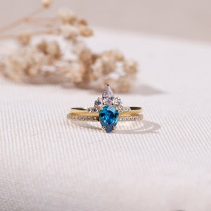 May include: A gold ring with a blue pear-shaped gemstone surrounded by smaller white gemstones. The ring is set on a white background.