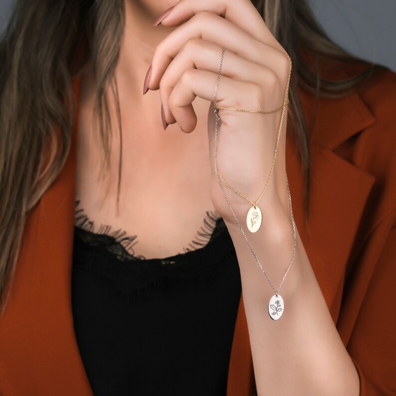 May include: Two layered necklaces with oval pendants. The top necklace is gold, and the bottom is silver. Each pendant features a floral design. The necklaces are displayed against a background of a person wearing a black top and a rust-colored blazer.