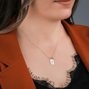 May include: A silver necklace with a delicate chain and an oval pendant. The pendant features a floral design and a small blue gemstone. The necklace is worn against a dark top and a rust-colored blazer.