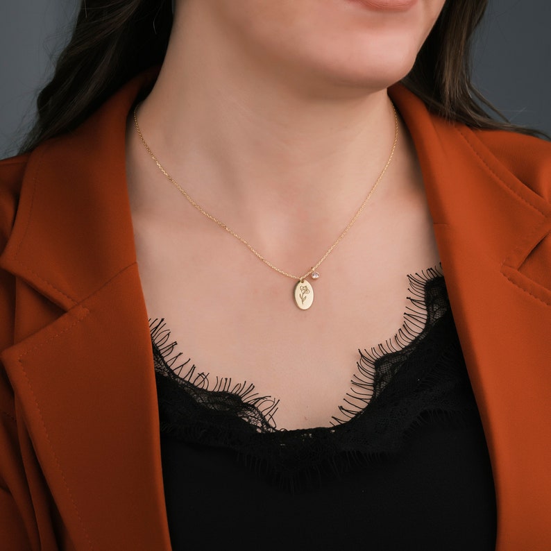 May include: A gold-colored necklace with a delicate chain. The pendant is an oval shape with a floral design and a small gemstone. The necklace is worn over a black top and a rust-colored blazer.