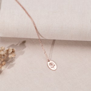 May include: Rose gold necklace featuring a delicate chain and an oval pendant. The pendant showcases a detailed rose illustration. The necklace is displayed on a neutral-toned fabric, highlighting its elegant design. A subtle, floral accent is visible in the corner.