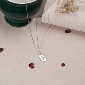May include: A silver necklace with a delicate chain and an oval pendant. The pendant features an engraved floral design. The necklace is displayed on a neutral-toned surface with dried flowers and red berries.
