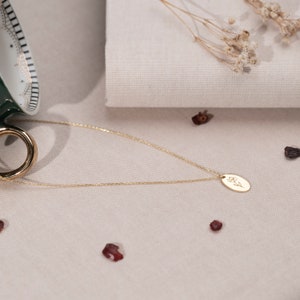 May include: A delicate gold necklace with a small oval pendant featuring a floral design. The necklace is displayed on a neutral-toned surface, with a gold ring and decorative elements in the background. The pendant's design is a simple, elegant floral motif.