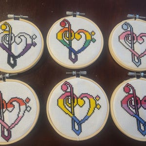May include: Six cross-stitch embroidery hoops featuring a treble clef and heart design. Each hoop has a different color scheme, including rainbow, pink, and purple, set against a white background. The hoops are displayed on a wooden surface.