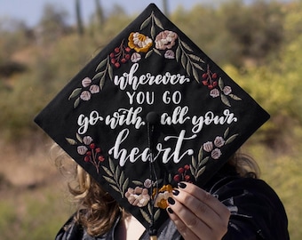 May include: Black graduation cap with white embroidery that reads "wherever you go go with all your heart". The cap is decorated with embroidered flowers in shades of red, pink, and yellow.