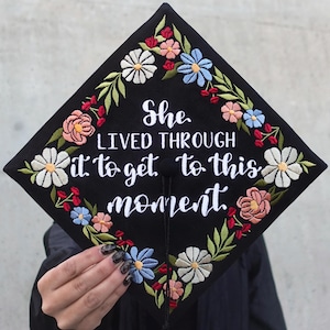 Könnte beinhalten: Schwarze Abschluss-Kappe mit einem floralen Stickmuster. Der Text "She lived through it to get to this moment" ist in Weiß auf die Kappe gestickt.