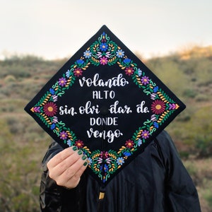 May include: A black graduation cap decorated with colorful embroidered flowers. The cap has the text "volando alto sin olvidar de donde vengo" written in white.