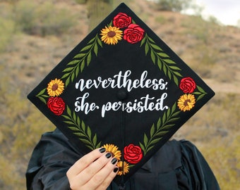 May include: A black graduation cap decorated with sunflowers, red roses, and green leaves. The cap has the text "nevertheless, she persisted" embroidered in white.