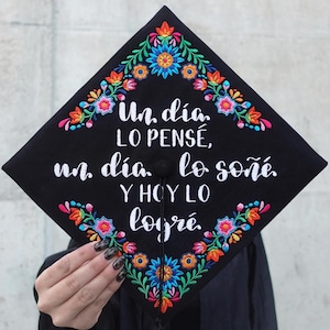 Könnte beinhalten: Eine schwarze Abschluss-Kappe, verziert mit bunten, gestickten Blumen und dem spanischen Text "Un día lo pensé, un día lo soñé y hoy lo logré."