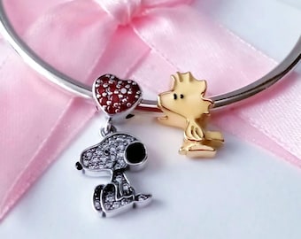 May include: A silver bracelet with three charms. One charm is a heart shape with red stones. Another charm is a silver Snoopy with clear stones. The third charm is a gold-colored Woodstock.