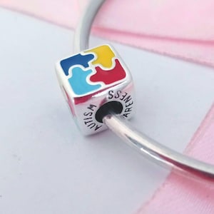 May include: A silver charm with a cube design featuring a colorful puzzle piece pattern in blue, yellow, and red. The charm has the words "AUTISM AWARENESS" engraved on its side. The charm is on a silver bracelet.