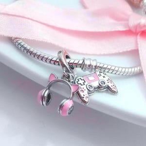 May include: A silver charm bracelet with three pink and silver charms. The charms include a pair of cat ear headphones, a video game controller, and a cross symbol. A pink ribbon is in the background.