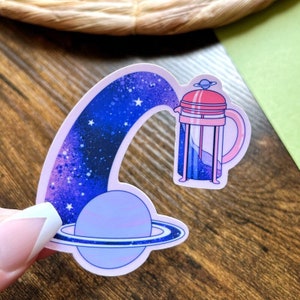 May include: A sticker of a pink and clear glass French press pouring coffee into a planet with rings. The background is a galaxy with stars and a purple and blue gradient.