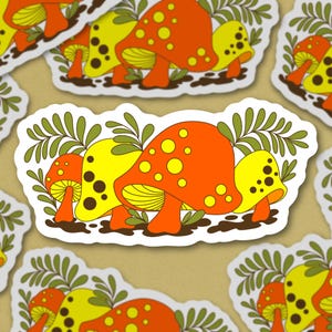May include: A sticker featuring three cartoon mushrooms with yellow and orange spots. The mushrooms are surrounded by green leaves.