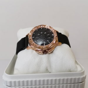 May include: A rose gold watch with a black mesh band and a black face with a decorative cutout design. The watch is in a white box.