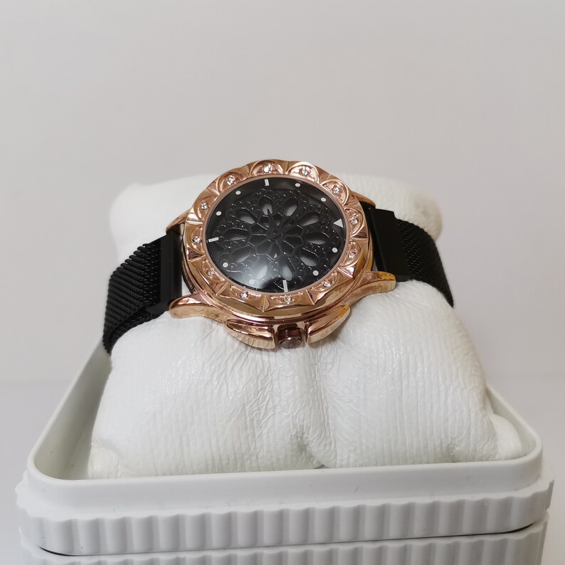 May include: A rose gold watch with a black mesh band and a black face with a decorative cutout design. The watch is in a white box.