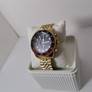 May include: Gold-toned wristwatch with a blue dial and red and black bezel. The watch features a gold link band and is displayed on a white cushion inside a white container. The watch has multiple sub-dials and gold hands.
