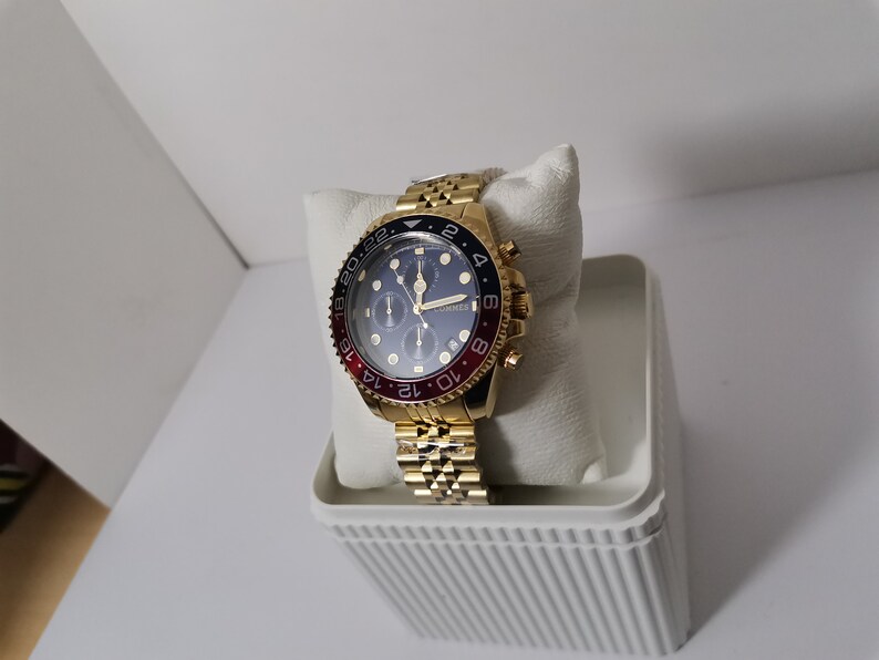 May include: Gold-toned wristwatch with a blue dial and red and black bezel. The watch features a gold link band and is displayed on a white cushion inside a white container. The watch has multiple sub-dials and gold hands.