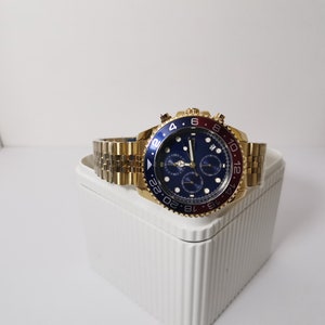 May include: A gold-toned wristwatch with a blue dial and a red and blue bezel. The watch has a gold-toned band and multiple sub-dials. The watch is displayed on a white surface.