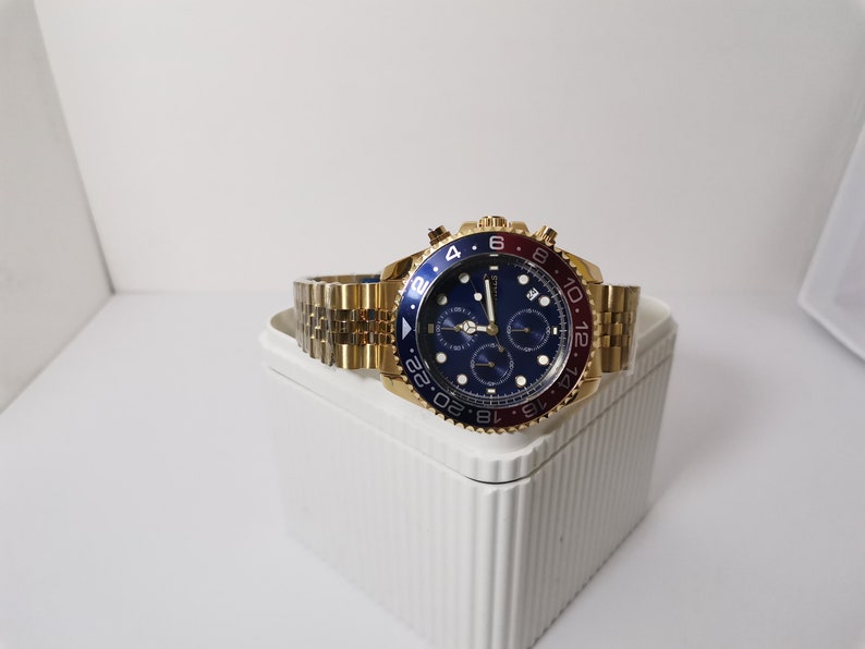 May include: A gold-toned wristwatch with a blue dial and a red and blue bezel. The watch has a gold-toned band and multiple sub-dials. The watch is displayed on a white surface.