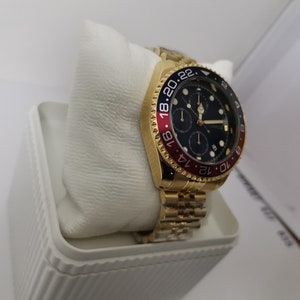 May include: A gold-toned wristwatch with a black dial and red and blue bezel. The watch features a gold band and is displayed on a white cushion inside a white box. The watch face has multiple sub-dials.