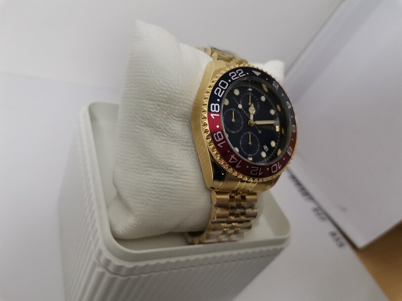 May include: A gold-toned wristwatch with a black dial and red and blue bezel. The watch features a gold band and is displayed on a white cushion inside a white box. The watch face has multiple sub-dials.