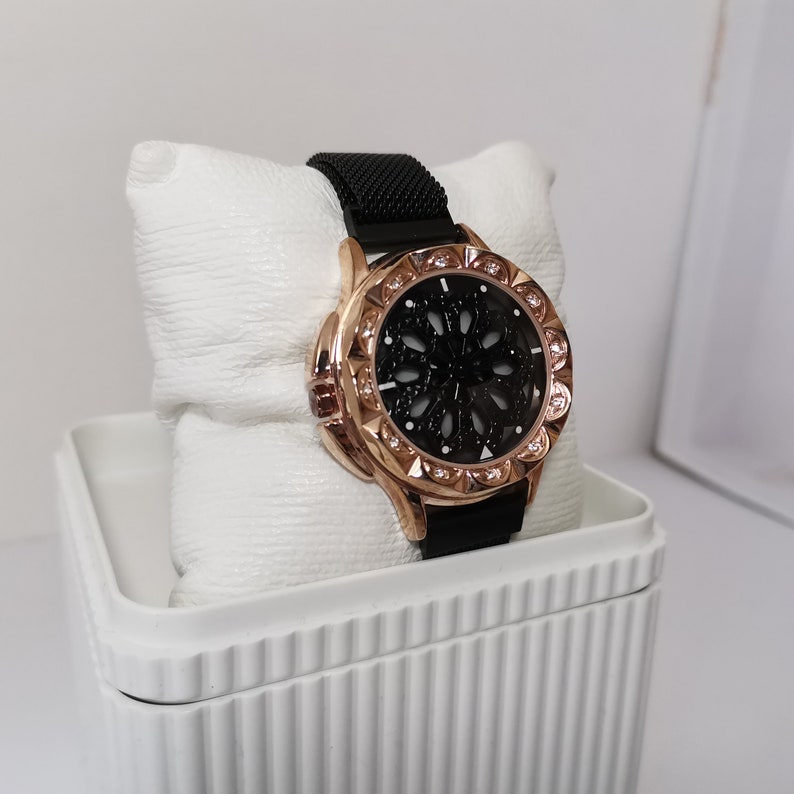 May include: A black and rose gold watch with a black face and a black mesh band. The watch is in a white box with a decorative pattern.