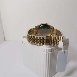 May include: A gold-toned wristwatch with a matching band is displayed on a white box. The watch's face is not visible, but the back shows a blue and silver design. The watch band is made of small, rectangular links.