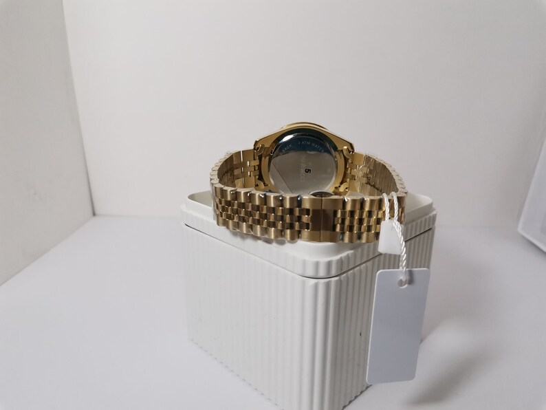 May include: A gold-toned wristwatch with a matching band is displayed on a white box. The watch's face is not visible, but the back shows a blue and silver design. The watch band is made of small, rectangular links.