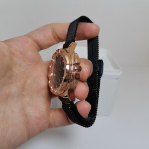 May include: A rose gold watch with a black mesh band. The watch face has a diamond-like design around the edge.