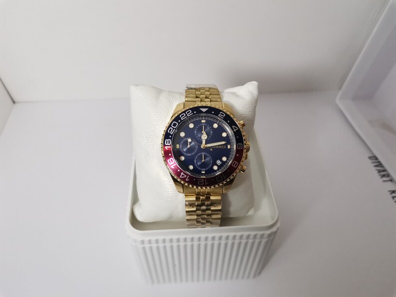 May include: A gold-toned wristwatch with a blue dial and red and blue bezel. The watch has a gold link band and is displayed on a white pillow inside a white box. The watch face has multiple sub-dials.