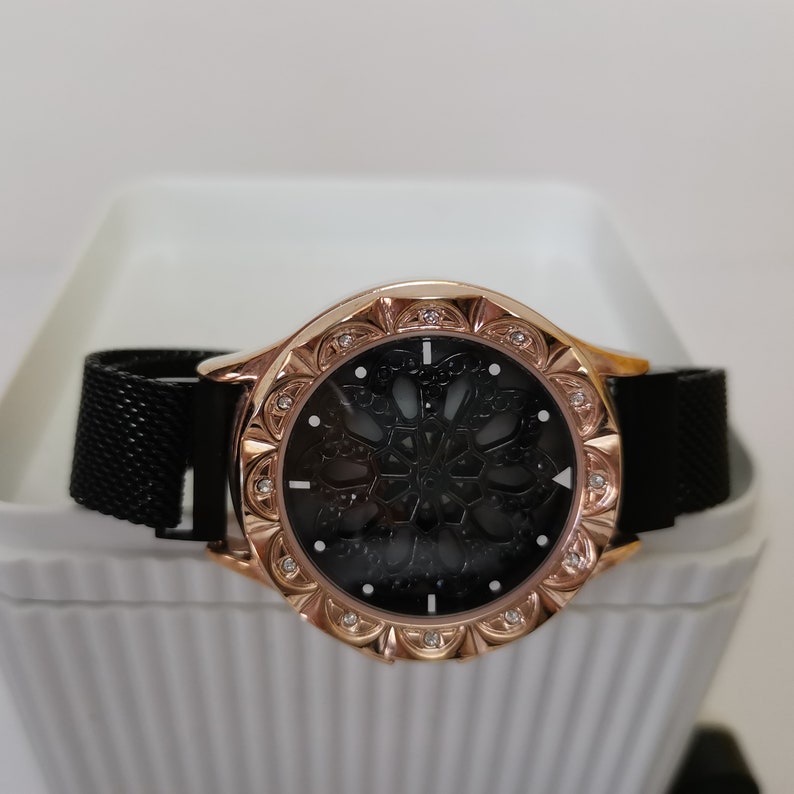 May include: A rose gold watch with a black mesh band and a black face with a decorative design. The watch has a round face and a bezel with small rhinestones.