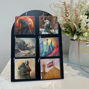 Può includere: Un espositore nero con sei stampe artistiche quadrate. Ogni stampa presenta un'illustrazione religiosa con figure in varie pose, dipinta in toni caldi e freddi. Le stampe sono disposte in due file da tre.