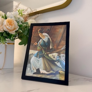 Può includere: Una stampa incorniciata raffigurante una scena storica con una figura in abito blu e bianco che brandisce una spada. L'opera d'arte è ambientata su uno sfondo di architettura in legno, con altre figure sullo sfondo. La cornice è nera.