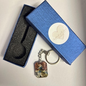 Può includere: Un portachiavi rettangolare con un'immagine religiosa, attaccato a un portachiavi argentato. Il portachiavi è esposto accanto a una scatola regalo blu con un adesivo bianco che dice "Handmade with love".