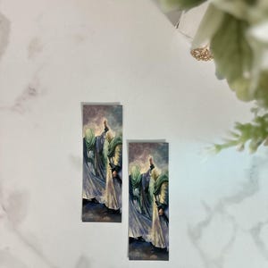 Può includere: Due segnalibri rettangolari con un dipinto di figure in abiti fluenti. Le figure hanno accenti verdi e oro, su uno sfondo di blu e viola tenui. I segnalibri sono su una superficie di marmo bianco.