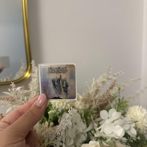 Può includere: Un piccolo adesivo quadrato con un'illustrazione di figure con abiti fluenti e scritte decorative. L'adesivo è tenuto davanti a un bouquet di fiori bianchi e color crema.