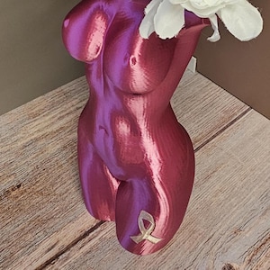 May include: A metallic pink vase in the shape of a female torso, holding white and purple flowers. The vase has a ribbon symbol on the lower part. The flowers are white and purple, with green leaves. The vase is on a wooden surface.