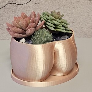 Peut inclure: Un pot en forme de fesses de couleur pêche avec une soucoupe assortie, contenant trois succulentes artificielles et de petits cailloux noirs. Le pot a une surface texturée et est conçu pour contenir de petites plantes. Les succulentes sont de différentes nuances de vert et de rose.