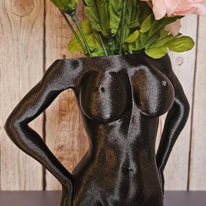 May include: A black, 3D printed vase shaped like a human torso. The vase is holding a bouquet of pink and purple flowers.